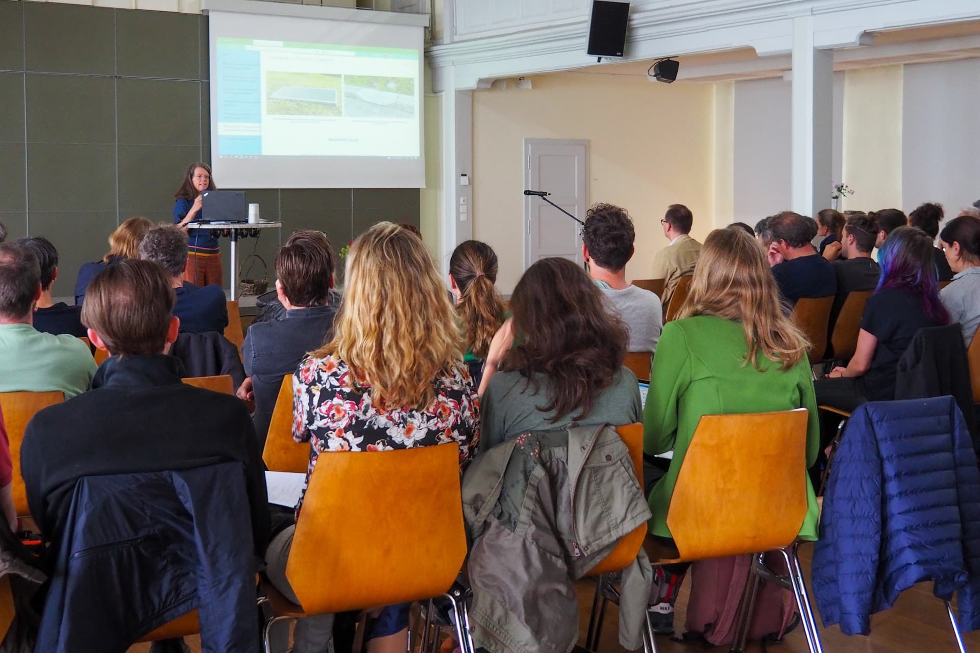 Environ 60 personnes sont assises dans une pièce et regardent un écran sur lequel quelque chose est présenté
