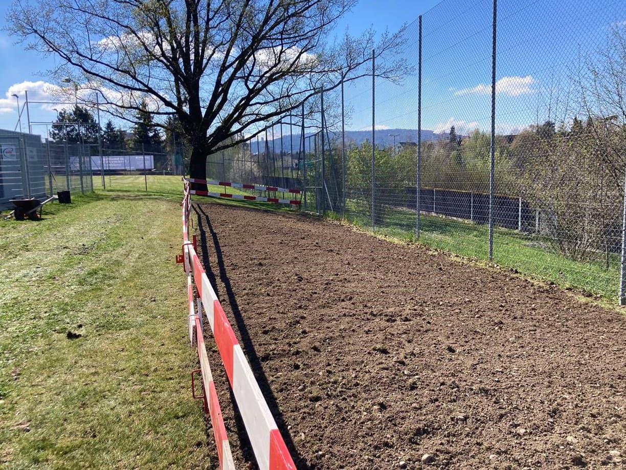 Surface prête à être ensemencée près d'un terrain de sport, clôturée par une barrière de chantier