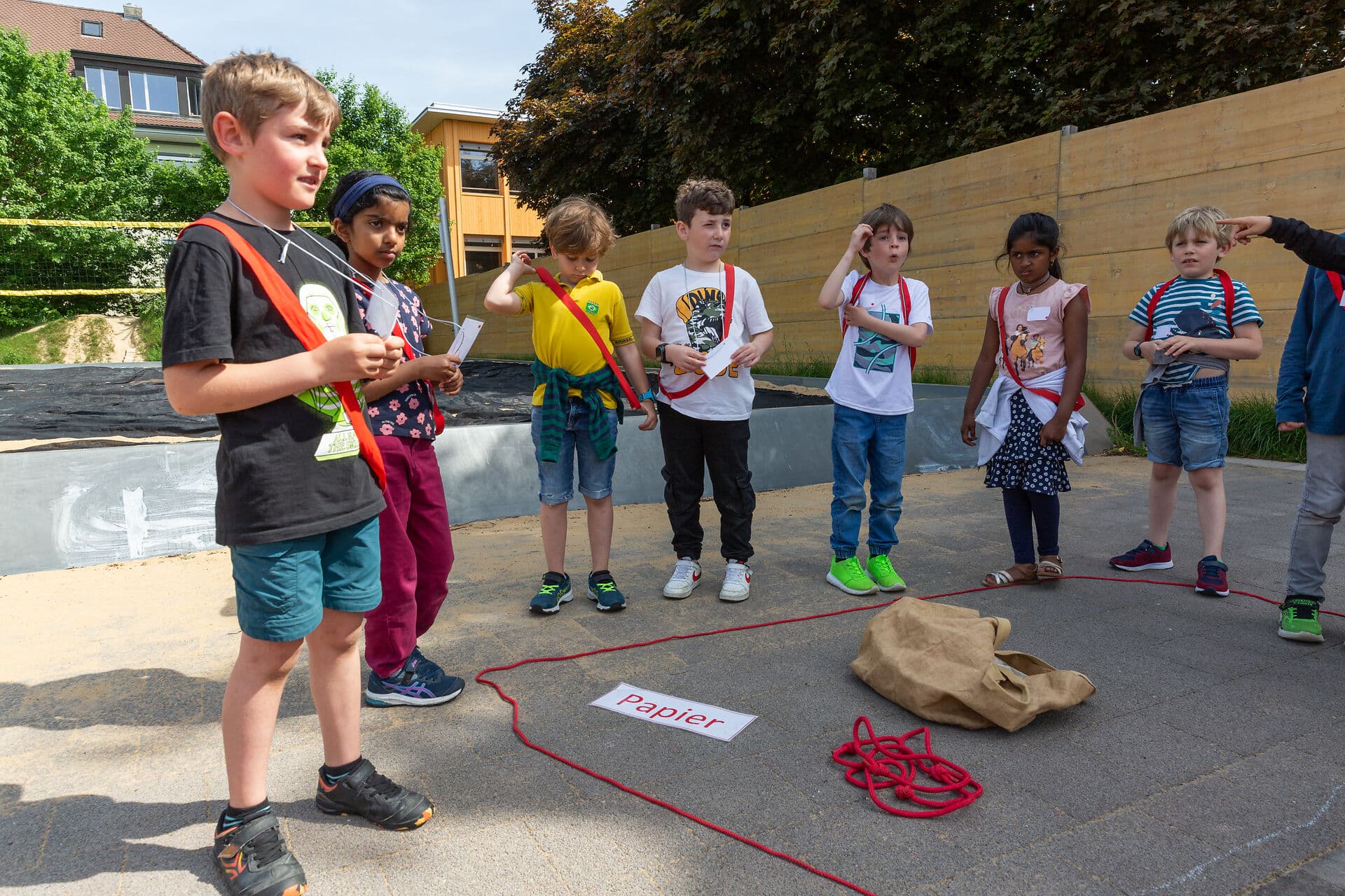 Kinder stehen im Halbkreis, vor ihnen am Boden eine braune Tasche, ein rotes Seil und ein Zettel mit Aufschrift «Papier»