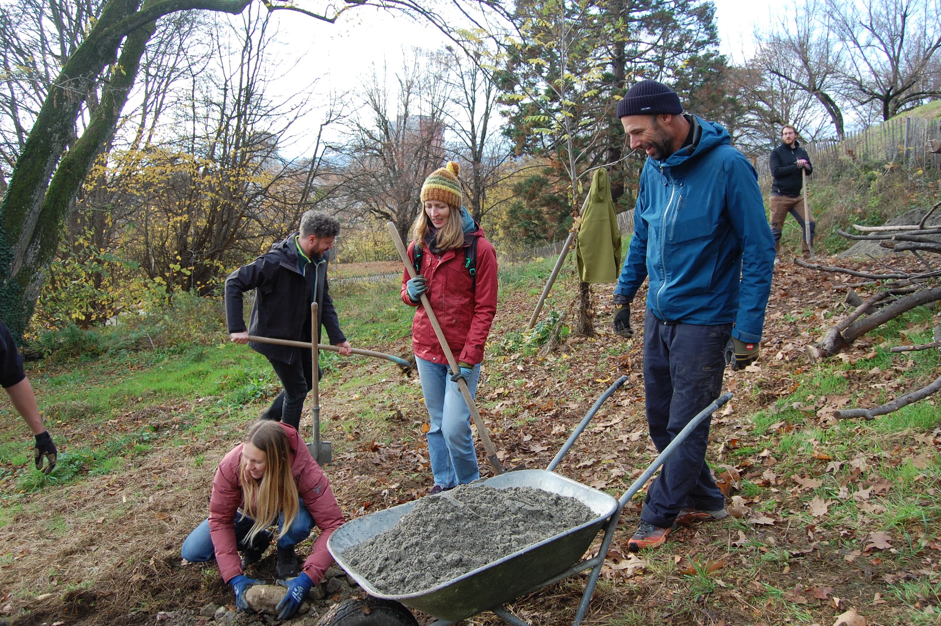 Des personnes manipulent des matériaux comme du sable, de la terre et des pierres avec une brouette et différents outils dans un parc arborisé.